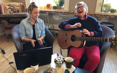 Two white people sit together on a couch. The person on the left is wearing a light blue denim jacket and is looking over at the person beside them. The other person is holding a guitar and is laughing.