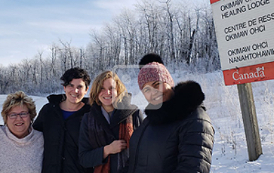 Four women stand side by side outdoors, in the winter. They are all wearing winter jackets, and one is wearing a touque. They are standing in front of a sign that reads "Okimaw Healing Lodge".