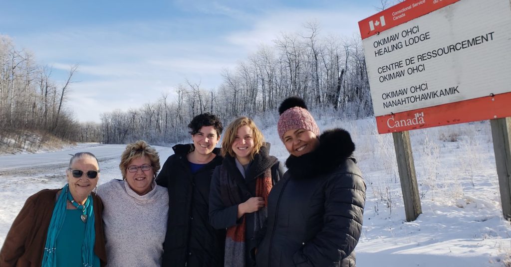 Five people stand outside during the winter, smiling at the camera. They are all wearing winter jackets, and are standing in front of a large sign that reads "Okimaw Ohci Healing Lodge. Centre De Ressourcement Okimaw Ohci. Okimaw Ohci Nanahtoahawkikamik."